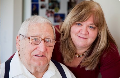 Elderly man with glasses and suspenders poses with a woman in a maroon top, both smiling warmly in a brightly lit room.