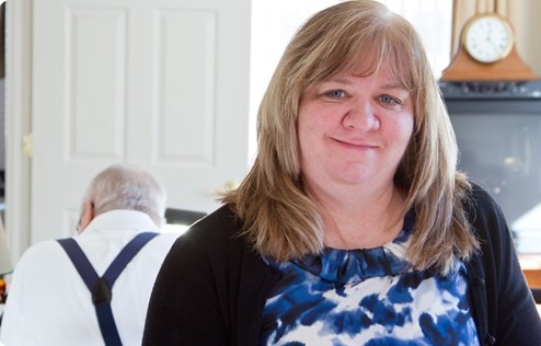 A woman with long hair and a blue-patterned blouse smiles at the camera, while an older man in the background focuses on a task.
