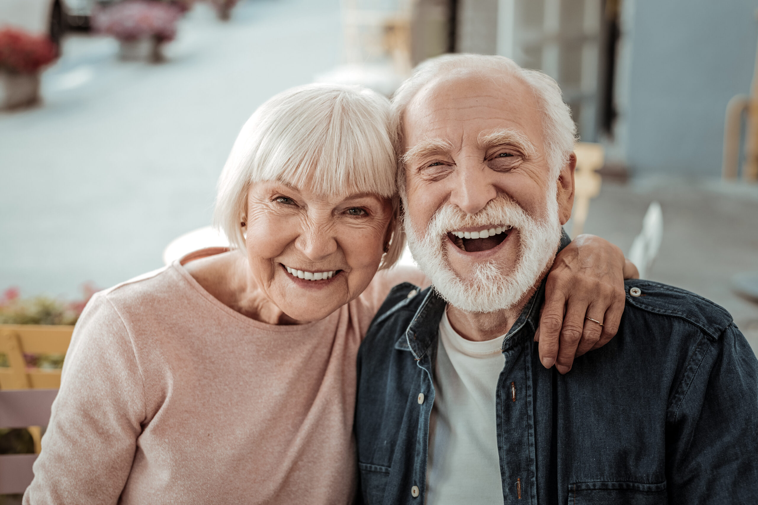 A cheerful older couple smiles at the camera, showcasing their close bond. Their joyful expressions convey happiness, set against a casual outdoor café background.