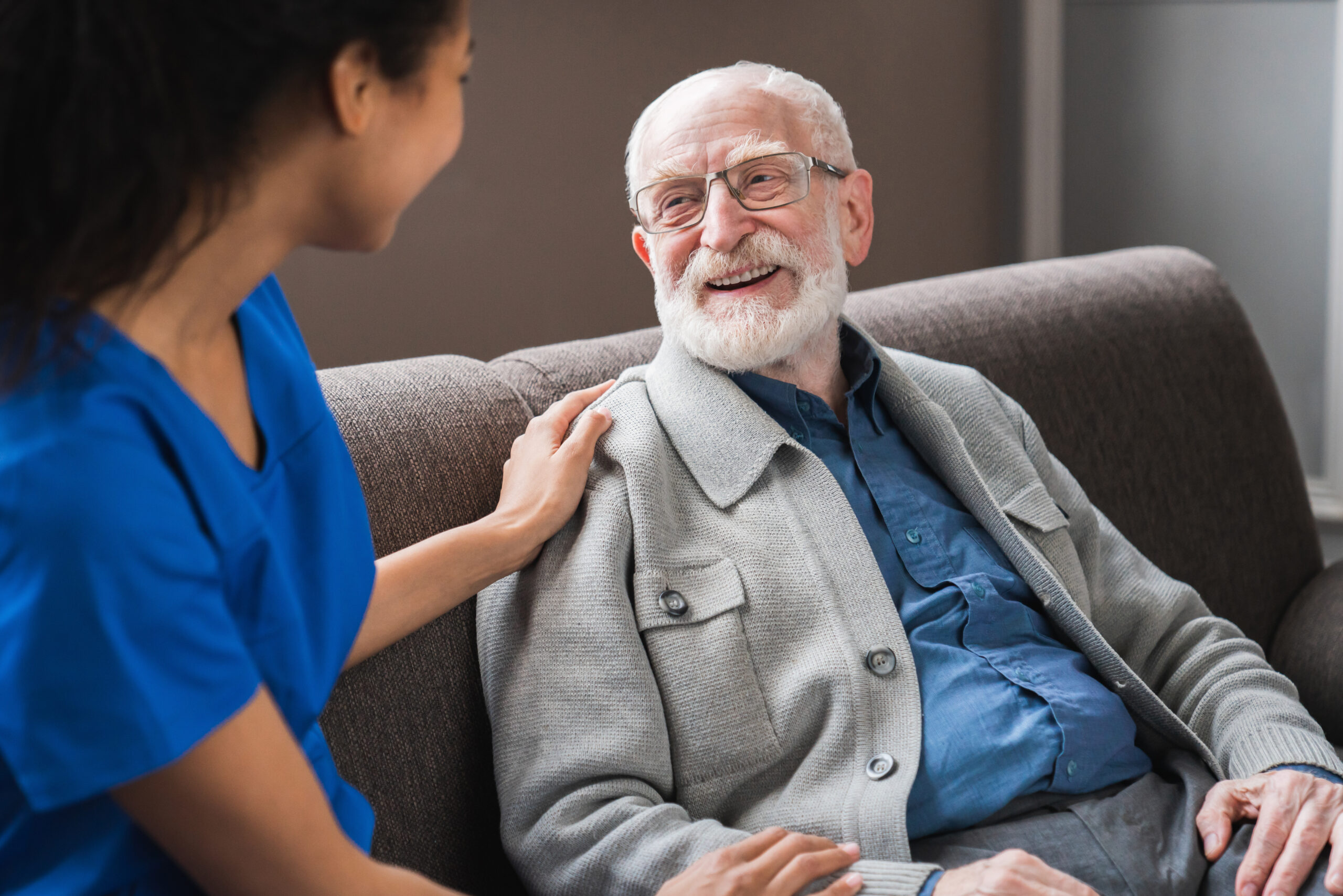 A caregiver in blue scrubs smiles at an elderly man with glasses, both engaged in a warm conversation while seated on a sofa.
