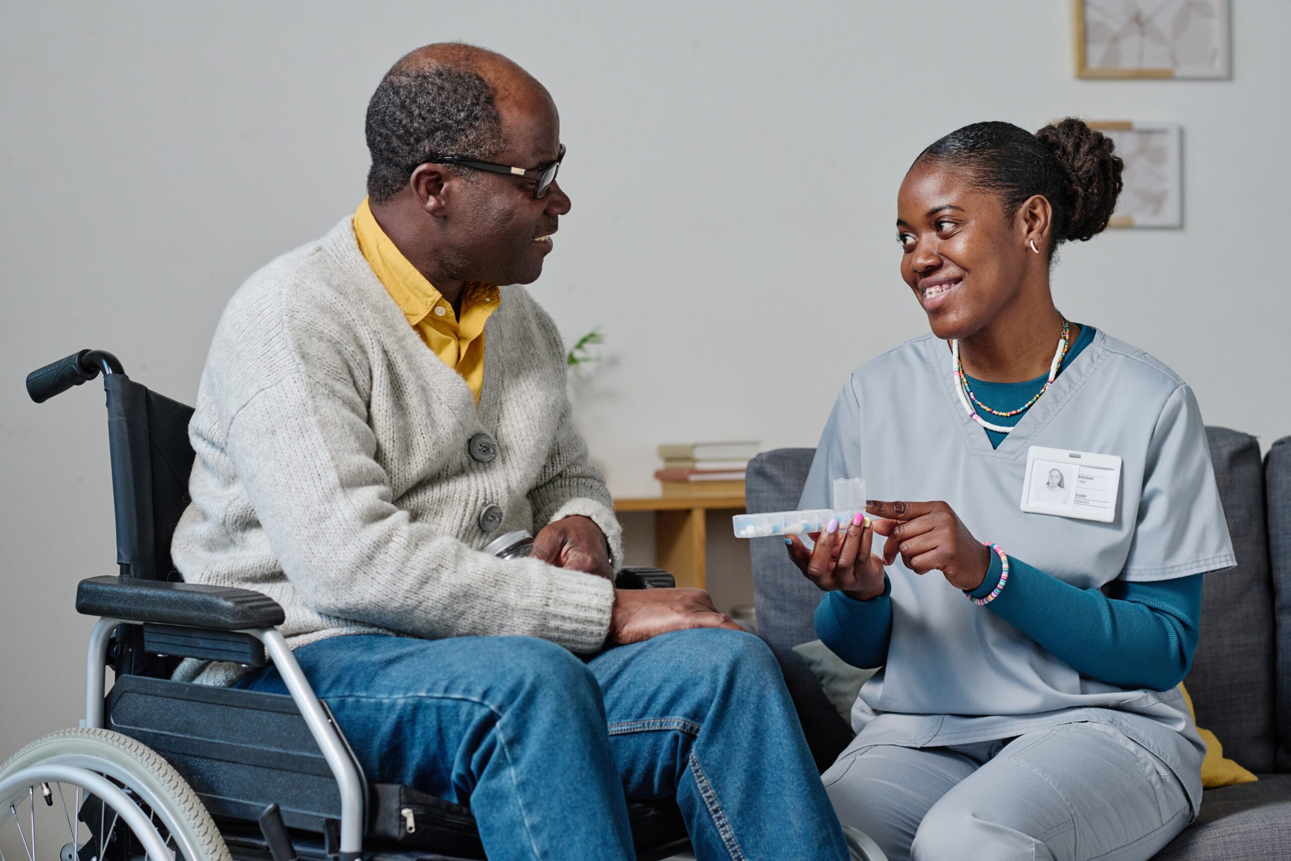 A caregiver in scrubs smiles while discussing medication management with a middle-aged man in a wheelchair, fostering a supportive healthcare environment.