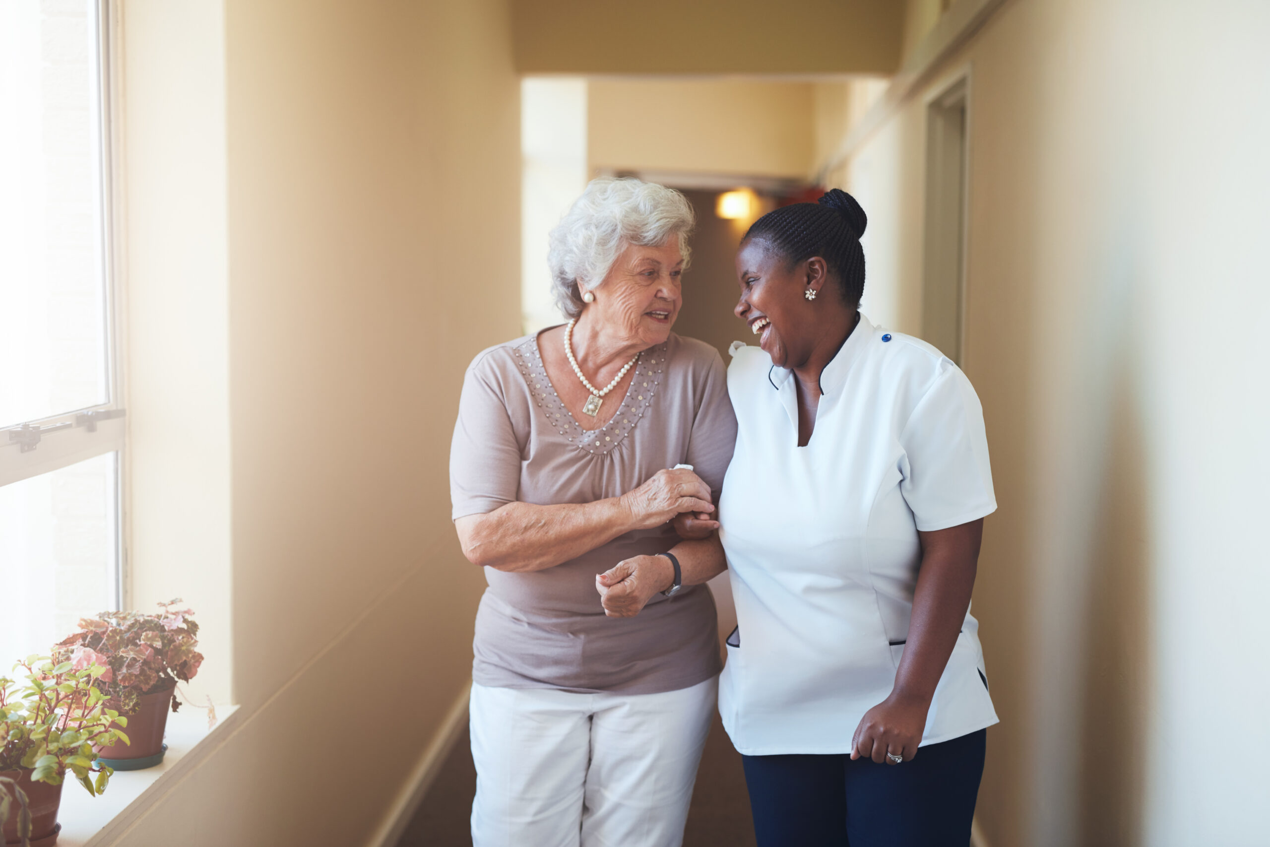A caregiver smiles and assists an elderly woman as they walk together down a well-lit hallway, highlighting companionship and support in healthcare.