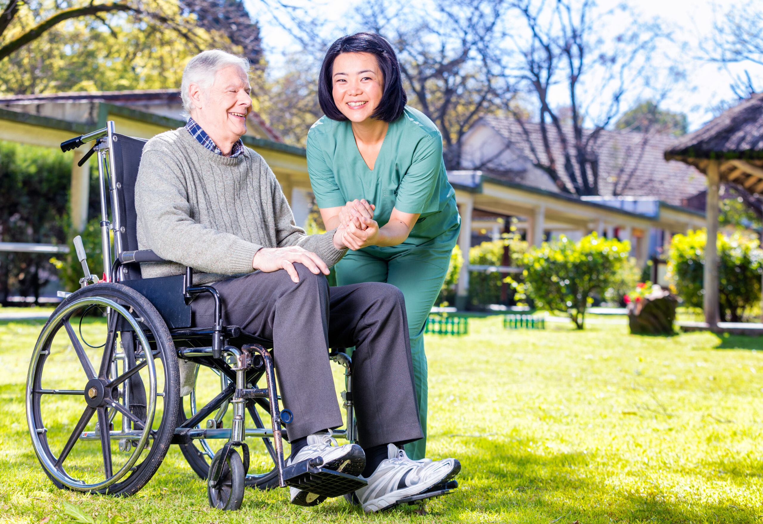 A smiling caregiver helps an elderly man in a wheelchair outdoors, emphasizing compassion and support in a healthcare setting.